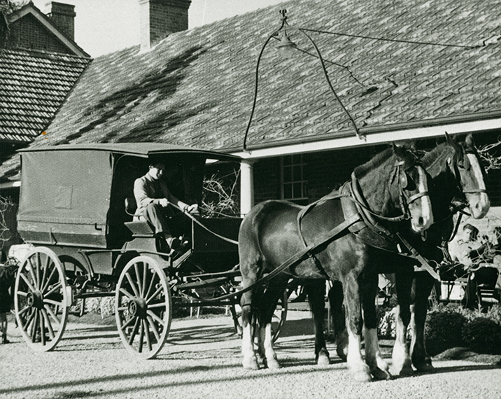 Student in a drag, harnessed to two horses, outside the Main Building - another student sitting on the verandah [Hawkesbury Agricultural College (HAC)]