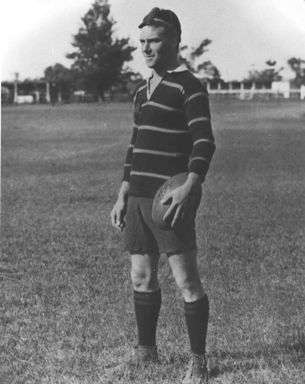 Student standing on a football oval, holding a football and wearing a striped rugby football jersey and honour cap [Hawkesbury Agricultural College (HAC)]