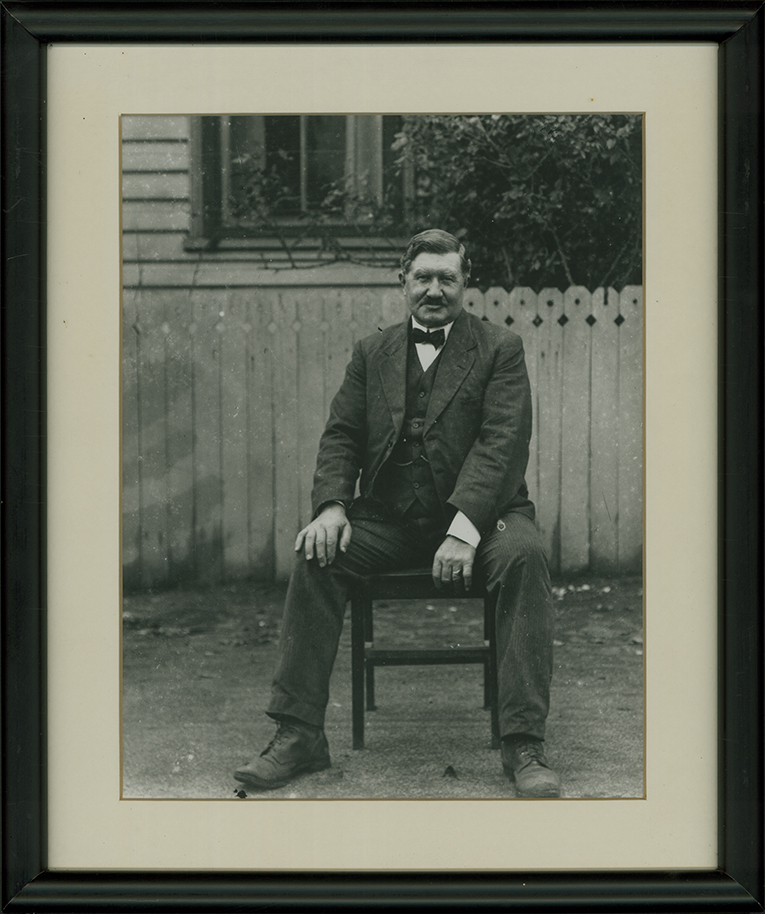 Suited man seated in front of fence [Hawkesbury Agricultural College (HAC)]