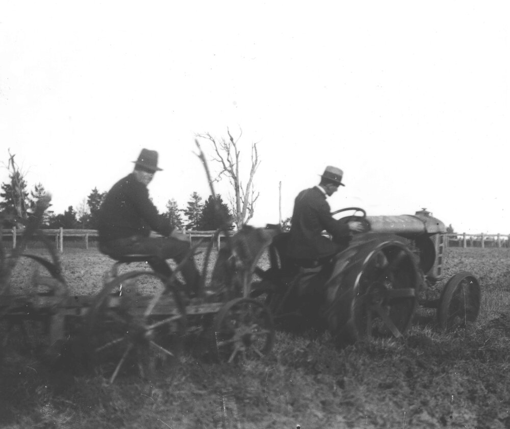A student under instruction, driving an early tractor pulling a plough, with the instructor sitting on the plough [Hawkesbury Agricultural College (HAC)]