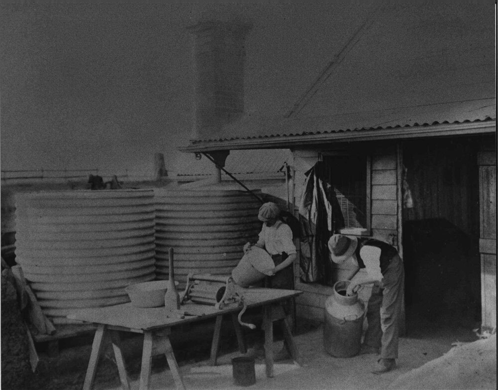 Two students cleaning milking buckets and cans outside the dairy (?) - there are two corrugated water tanks to one side [Hawkesbury Agricultural College (HAC)]