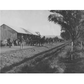 Teams of horses passing a large farm shed, setting out for work in the fields - two tractors parked by the side of the dirt road [Hawkesbury Agricultural College (HAC)]