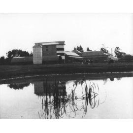 Twin silos with underground silo, and other dairy buildings behind, dam in the foreground with cows in field in front of buildings [Hawkesbury Agricultural College (HAC)]