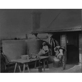 Two students cleaning milking buckets and cans outside the dairy (?) - there are two corrugated water tanks to one side [Hawkesbury Agricultural College (HAC)]